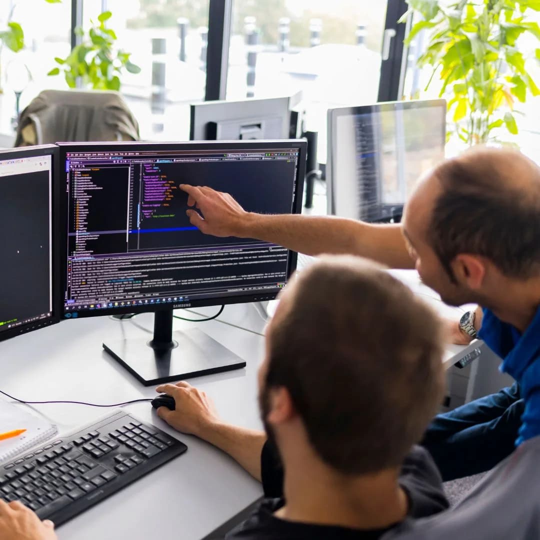 Zwei Entwickler am Schreibtisch, einer zeigt auf den Code am Monitor